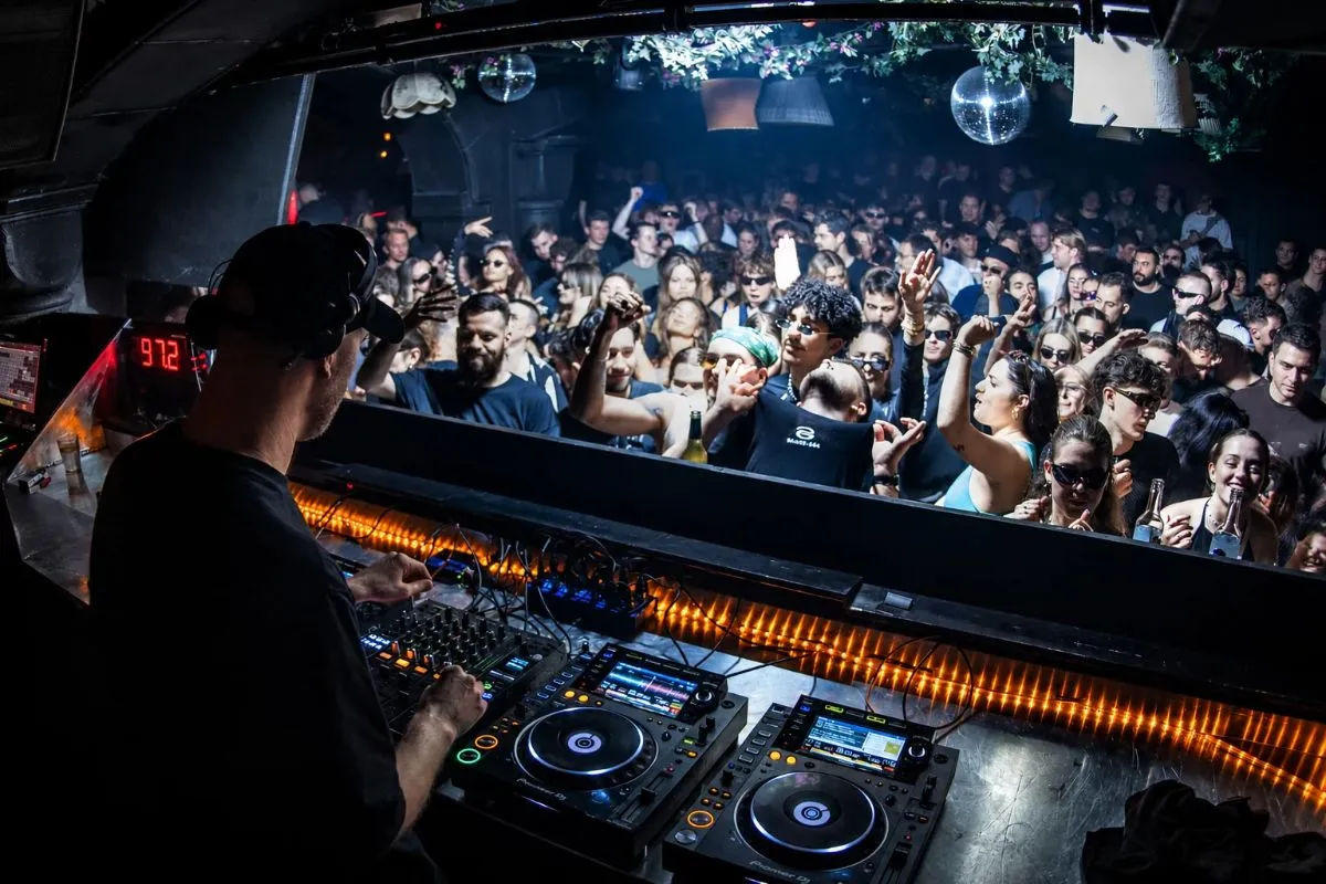 DJ seleccionando música frente a una pista de baile mientras una pantalla muestra información de los tracks.